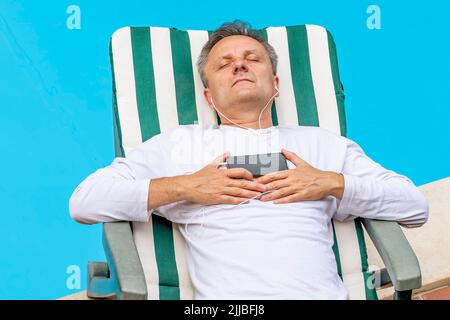 Ein weißer Mann mittleren Alters entspannt sich, indem er Musik mit Kopfhörern von seinem Smartphone am Pool hört Stockfoto