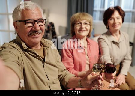 Glücklicher älterer Mann in Brillen, der mit Gläsern Wein von zwei älteren Frauen zusammenklappte, während er eine Heimparty oder ein Treffen mit Freunden genoss Stockfoto