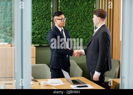 Seitenansicht eines Porträts von zwei Geschäftspartnern, die sich die Hände schütteln, während sie sich im Konferenzraum mit umweltfreundlichem Design und Kopierfläche treffen Stockfoto