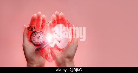 Embryo Silhouette aus Papier und Uhr in Frauenhand mit gebrochenem Herzen. Hände auf der linken Seite. Rosafarbener Boden mit Kopierfläche. Stockfoto