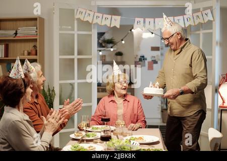 Glücklicher älterer Mann, der hausgemachten Geburtstagskuchen für seine Frau und ihre Freunde in Mützen trägt, die am Tisch auf der Party serviert werden Stockfoto