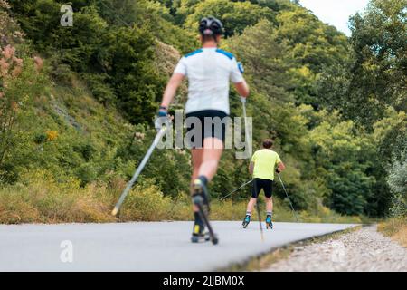 Zwei sportliche Männer trainieren auf dem Rollski auf der Landstraße, Rückansicht. Unschärfer Sportler im Vordergrund. Low-Angle-Ansicht. Konzept der Sportwettbewerbe Stockfoto