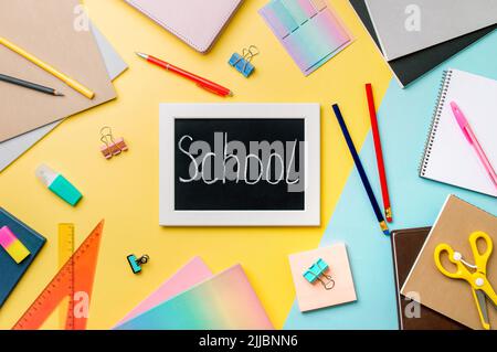 Tafel mit Briefen Schule mit Kreide gemacht. Schulbedarf, Bleistifte, Scheren und farbige Bücher auf gelbem und blauem Hintergrund. Ansicht von oben oder flach liegend. Zurück zum Schulkonzept Stockfoto