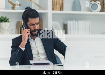 Ein hübscher Mann, der in seinem Büro mit weißem Design telefoniert, während er an einem Laptop arbeitet. Stockfoto