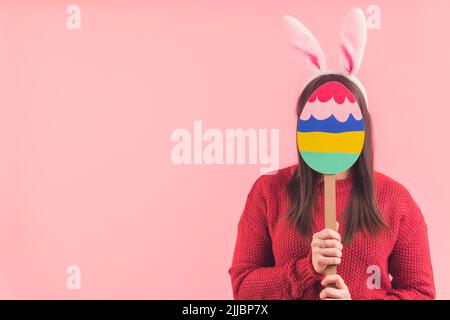 Brünette Frau hält bemaltes osterei vor ihrem Gesicht trägt Hasenohren und roten Pullover. Rosafarbene Studioaufnahme. Hochwertige Fotos Stockfoto
