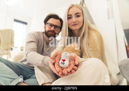 Weiß erwartenden Paar mit gestrickten Baby-Schuh Ich liebe Vater Blick auf die Kamera im Schlafzimmer sitzen. Fokus auf Vordergrund. Innenaufnahme. Hochwertige Fotos Stockfoto