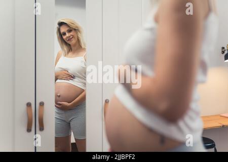 Kaukasische junge Erwachsene blonde Schwangeren Frau Blick auf ihren großen Bauch in der Garderobe Spiegel. Innenaufnahme. Konzentrieren Sie sich auf den Hintergrund. Hochwertige Fotos Stockfoto