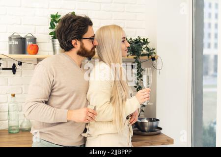 Junge weiße Paar Brünette Mann mit einem Bart und Brille hält blonde Frau in der Küche trinken Champagner durch das Fenster schauen. Innenaufnahme. Hochwertige Fotos Stockfoto