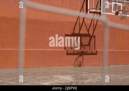 Blick aus dem niedrigen Winkel des Outdoor Basketball Backboards an einem sonnigen Tag, Schatten reflektiert auf orange Wand. Städtischer Sportplatz.verschwommene Nahaufnahme des Zauns. Stockfoto