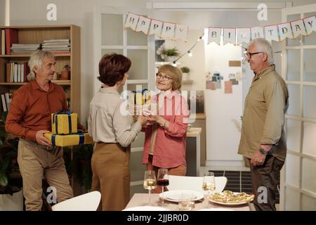 Glückliches reifes Paar, das verpackte und verpackte geschenkkartons mit Geburtstagsgeschenken an lächelnde, gealterte Frau gibt, die am geservierten Tisch steht Stockfoto
