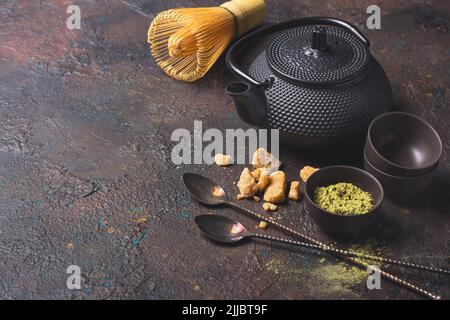 Bio-grünes Matcha-Teepulver in Schüsseln oder Tassen mit Bambusbesen, Vintage-Löffeln, Rohrzucker und schwarzem asiatischen Wasserkocher auf dunklem Beton-Hintergrund Stockfoto