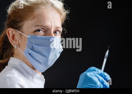 Eine junge Ärztin mit einer Spritze schaut auf die Kamera. Eine Krankenschwester, die eine medizinische Maske und blaue Latexhandschuhe trägt. Medizin und Gesundheitswesen. Impfung Stockfoto