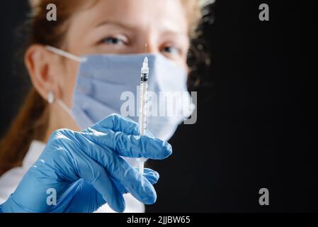 Eine junge Ärztin mit einer Spritze schaut auf die Kamera. Eine Krankenschwester, die eine medizinische Maske und blaue Latexhandschuhe trägt. Medizin und Gesundheitswesen. Impfung Stockfoto