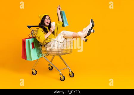 Weiß aufgeregt Frau lachen und mit Handy in Warenkorb isoliert über gelben Hintergrund Stockfoto