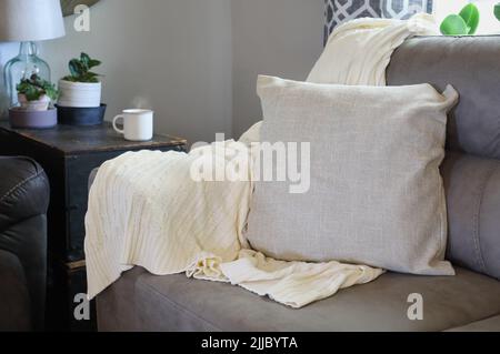 Weiche weiße Strickdecke über den Arm einer grauen Couch in einem Wohnzimmer im Landhausstil. Selektiver Fokus mit unscharfem Hintergrund. Stockfoto