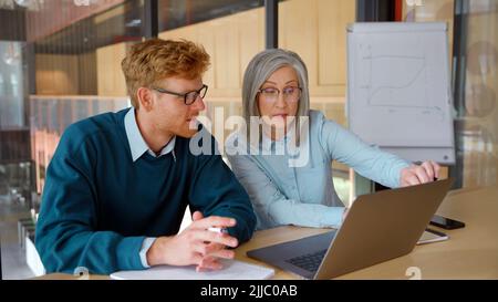 Reifer Mentor, der Praktikant unterrichtet und die Online-Arbeit mit einem Laptop im Büro erklärt. Stockfoto