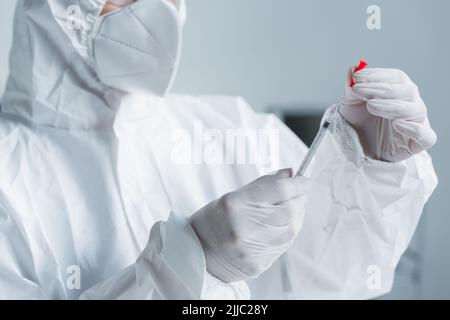 Beschnittene Ansicht des Wissenschaftlers in einem Hazmat-Anzug mit Latexhandschuhen, die die Spritze im Labor halten Stockfoto