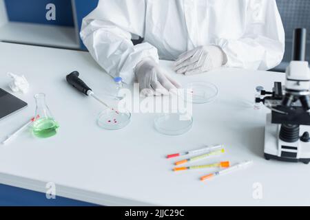 Beschnittene Ansicht eines Wissenschaftlers in einem Hazmat-Anzug, der Glas in der Nähe von Petrischalen und Mikroskop im Labor hält Stockfoto