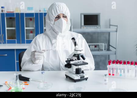 Wissenschaftler im Gefahrstoffanzug, der Glas in der Nähe des Mikroskops und der Reagenzgläser im Labor hält Stockfoto