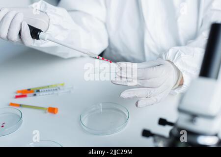 Beschnittene Ansicht des Wissenschaftlers in einem Hazmat-Anzug, der Glas und elektronische Pipetten im Labor hält Stockfoto