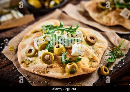 Von oben geschnittener Feta-Käse mit Oliven und Rucola auf knusprigem Pizzabrot, serviert auf Holzschneidebrett in der Restaurantküche Stockfoto