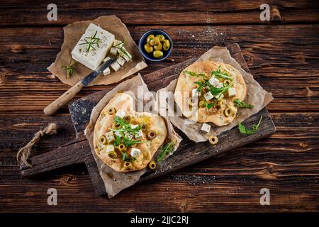 Blick von oben auf ein köstliches Gericht mit knusprigen Pita-Broten, garniert mit Feta-Käse-Oliven und Kräutern, serviert auf einem Holzbrett auf dem Tisch Stockfoto