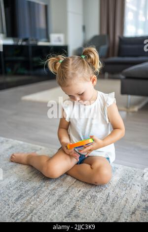 Nette kleine Mädchen spielen mit Begeisterung Pop it sensorische Spielzeug Kreis Form. Kleine weibliche Pressen bunte Regenbogen quishy weiche Silikon-Blasen zu Hause Stockfoto