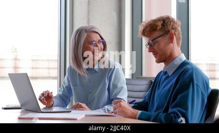 Reifer Mentor, der Praktikant unterrichtet und die Online-Arbeit mit einem Laptop im Büro erklärt. Stockfoto