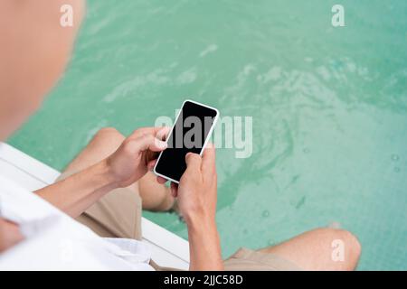 Beschnittene Ansicht von Mann Messaging auf dem Smartphone mit leerem Bildschirm in der Nähe von Pool mit türkisfarbenem Wasser Stockfoto