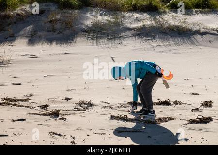 Eine junge Frau mit türkisfarbener Jacke geht am Strand entlang und sammelt Müll und Plastik, um bei ökologischen Aufgaben zu helfen Stockfoto