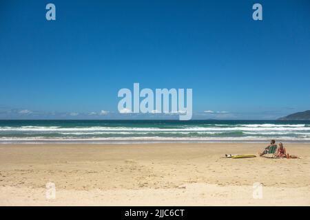 Ein paar Surfer genießen ein Sonnenbad, sitzen auf dem Sand in der Nähe von zwei Surfbrettern. Ruhiges Meer, blauer Himmel, sauberer Strand, schöner Ort, coole, entspannte Atmosphäre, Romantik, Stockfoto