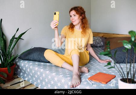 Das junge rothaarige Mädchen macht ein Selfie zu Hause auf dem Sofa auf einem Smartphone Stockfoto