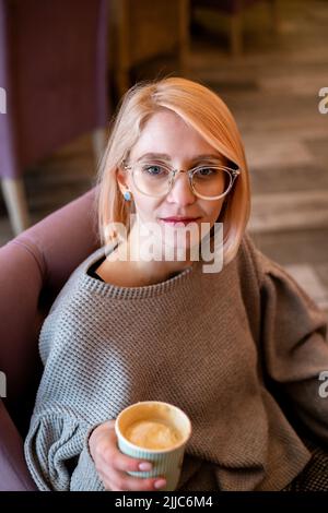 Junge attraktive blonde Mädchen im Café trinkt Kaffee aus einer Papiertasse und entspannt Stockfoto