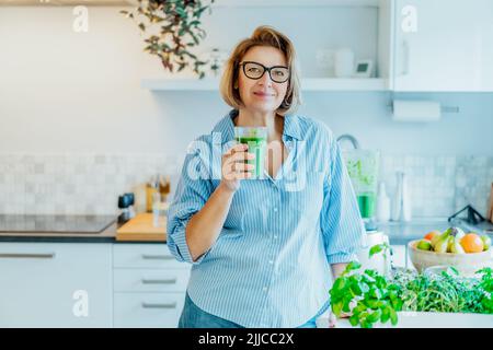 Lächelnde Frau mittleren Alters mit einem Glas Detox-Shake und grünem Smoothie in der Küche. Gesunde Ernährung, Essen, Kochen. Natürliche Anti-Aging erfüllt Stockfoto
