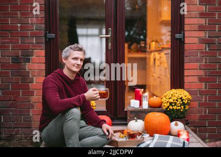 Porträt eines jungen Mannes mit heißem Tee auf der Veranda seines Hauses sitzen. Gemütliche Ideen, wie man Zeit zu Hause verbringt. Herbsttee im Freien auf dem Hauseingang Stockfoto