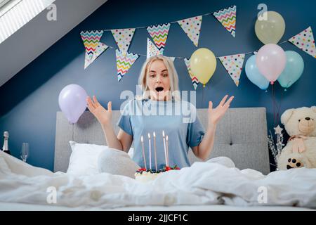 Fröhliche Frau in Pyjama und Partymütze, die Kerzen auf dem Geburtstagskuchen auf dem Bett im dekorierten Schlafzimmer ausblasen. Machen Sie einen Wunschprozess. Morgenüberraschung. Glücklich Stockfoto