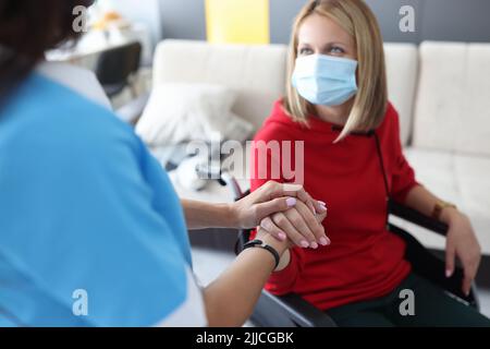Rehabilitationsarzt in schützender medizinischer Maske kommuniziert mit einer behinderten Frau im Rollstuhl Stockfoto