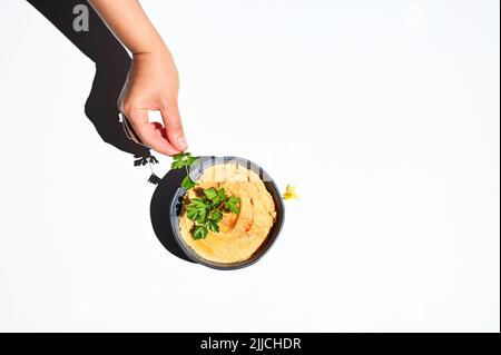 Draufsicht auf eine Hand, die Petersilie neben einer Schüssel mit Hummus hält, isoliert auf weißem Hintergrund mit Platz für Text Stockfoto