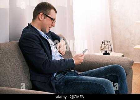 Portrait Seitenansicht des kaukasischen Mannes mit Jacke und Brille, der in offener Position auf dem Sofa sitzt und vor einem aufklappbaren Fenster mit Vorhang telefoniert Stockfoto