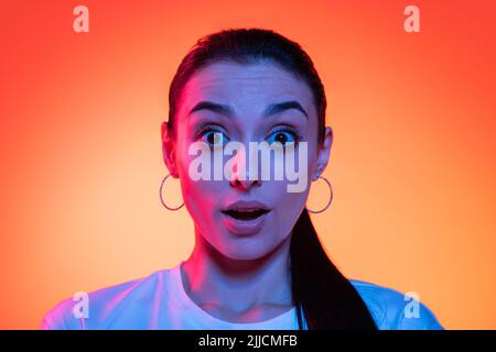 Nahaufnahme Gesicht von jungen attraktiven Mädchen Blick auf Kamera isoliert auf orangenen Hintergrund in Neonlicht. Konzept der Emotionen, Mimik Stockfoto