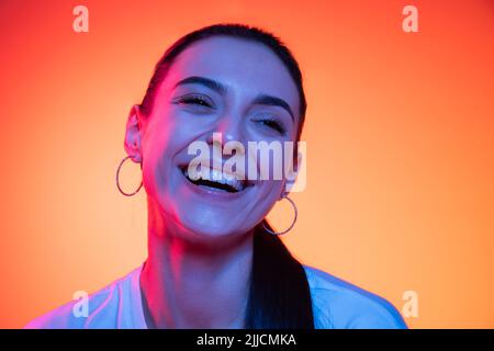 Nahaufnahme Gesicht von jungen attraktiven Mädchen Blick auf Kamera isoliert auf orangenen Hintergrund in Neonlicht. Konzept der Emotionen, Mimik Stockfoto