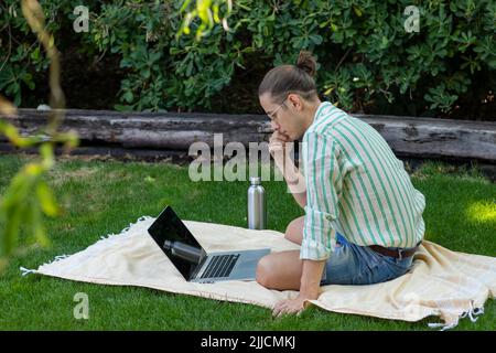 Seitenansicht des jungen Mannes, der im Park auf einem Handtuch sitzt, konzentrierte sich im Frühjahr auf die Arbeit an seinem neuen Projekt rund um die Welt Stockfoto