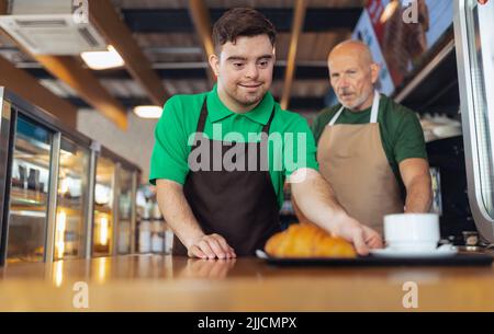 Glücklicher Kellner mit Down-Syndrom, der Kaffee mit Hilfe seines Kollegen im Café serviert. Stockfoto