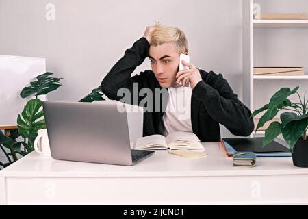 Ein junger, frustrierter, schockierter Mann hält sich am Kopf und starrt mit weit geöffneten Augen auf einen Laptop-Bildschirm, während er an seinem Schreibtisch im Büro anruft Stockfoto