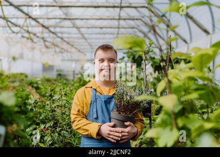 Ein glücklicher junger Mitarbeiter mit Down-Syndrom, der im Gartencenter arbeitet und sich um Blumen kümmert. Stockfoto