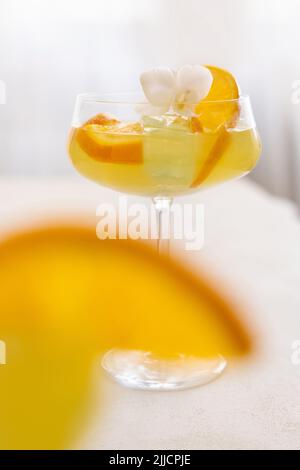 Transparenter Cocktail mit Eiswürfeln in einem Champagner-Coupé-Glas, verziert mit Orangenscheiben und weißen Orchideenblüten aus nächster Nähe. Tropisches Getränk im Stockfoto