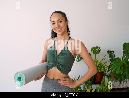 Eine junge, multiethnische Frau steht mit ihrer Yogamatte im pflanzengefüllten Studio Stockfoto