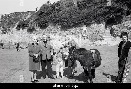1970s, historische, Menschen, die für ihr Foto stehen, neben der Anmietung von Eseln an einem Strand, England, Großbritannien. Der Bursche, der die Esel anheuert, steht dort. Stockfoto