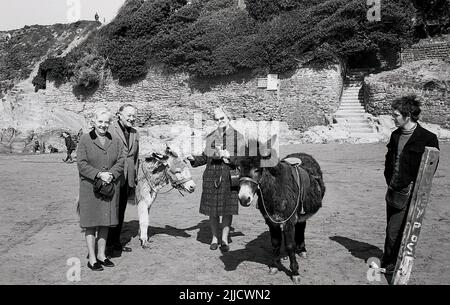1970s, historische, Menschen, die für ihr Foto stehen, neben der Anmietung von Eseln an einem Strand, England, Großbritannien. Der Bursche, der die Esel anheuert, steht dort. Stockfoto