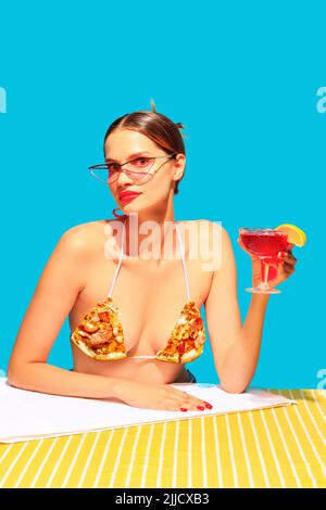 Stilvolle junge Frau trinkt leckeren Cocktail isoliert auf blauem Hintergrund. Pizza Badeanzug Element Stockfoto